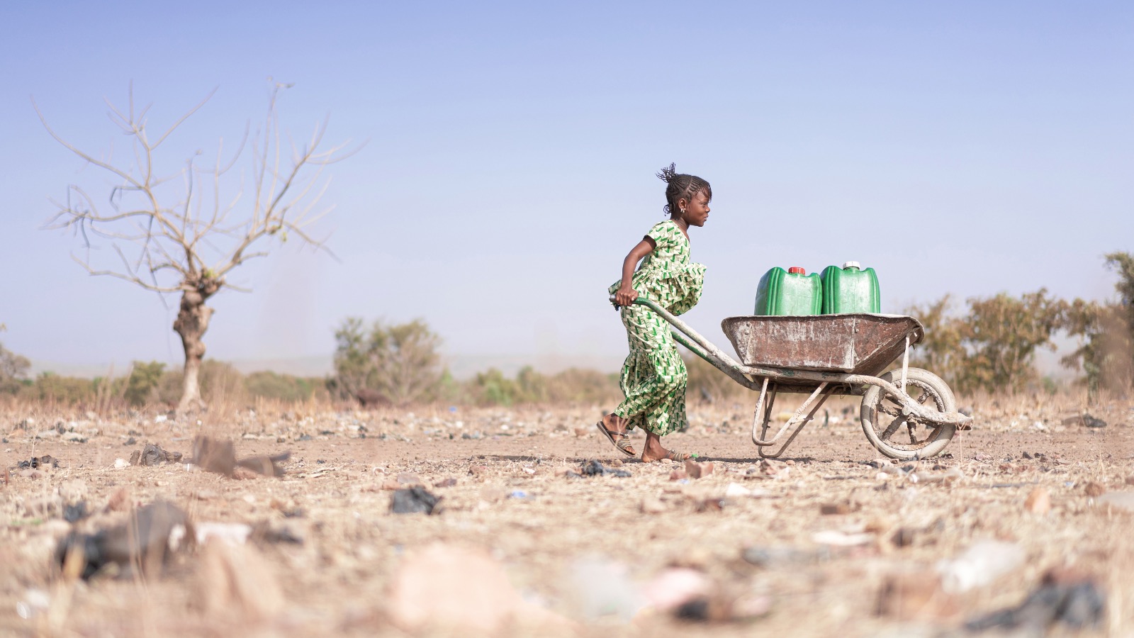 A young child carries a wheelbarrow with two water cans across a parched landscape.