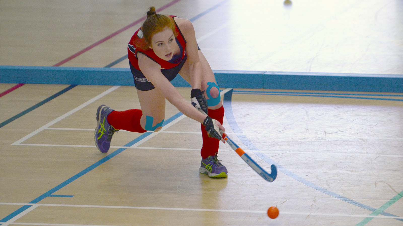 A female hockey player takes a swing at the ball on an indoor court.