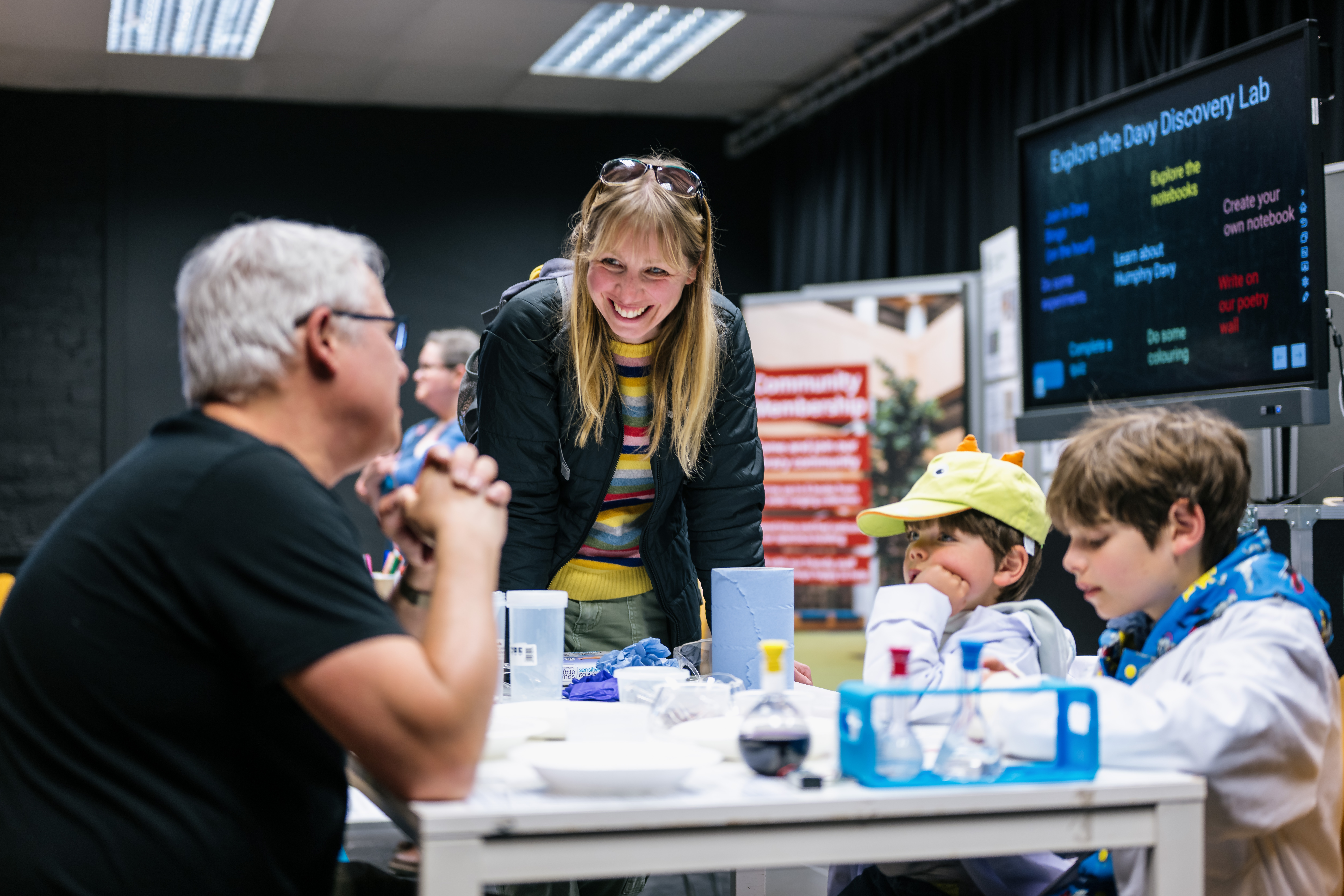 two children in lab coats sit at a table and a parent listen to information from a member of staff