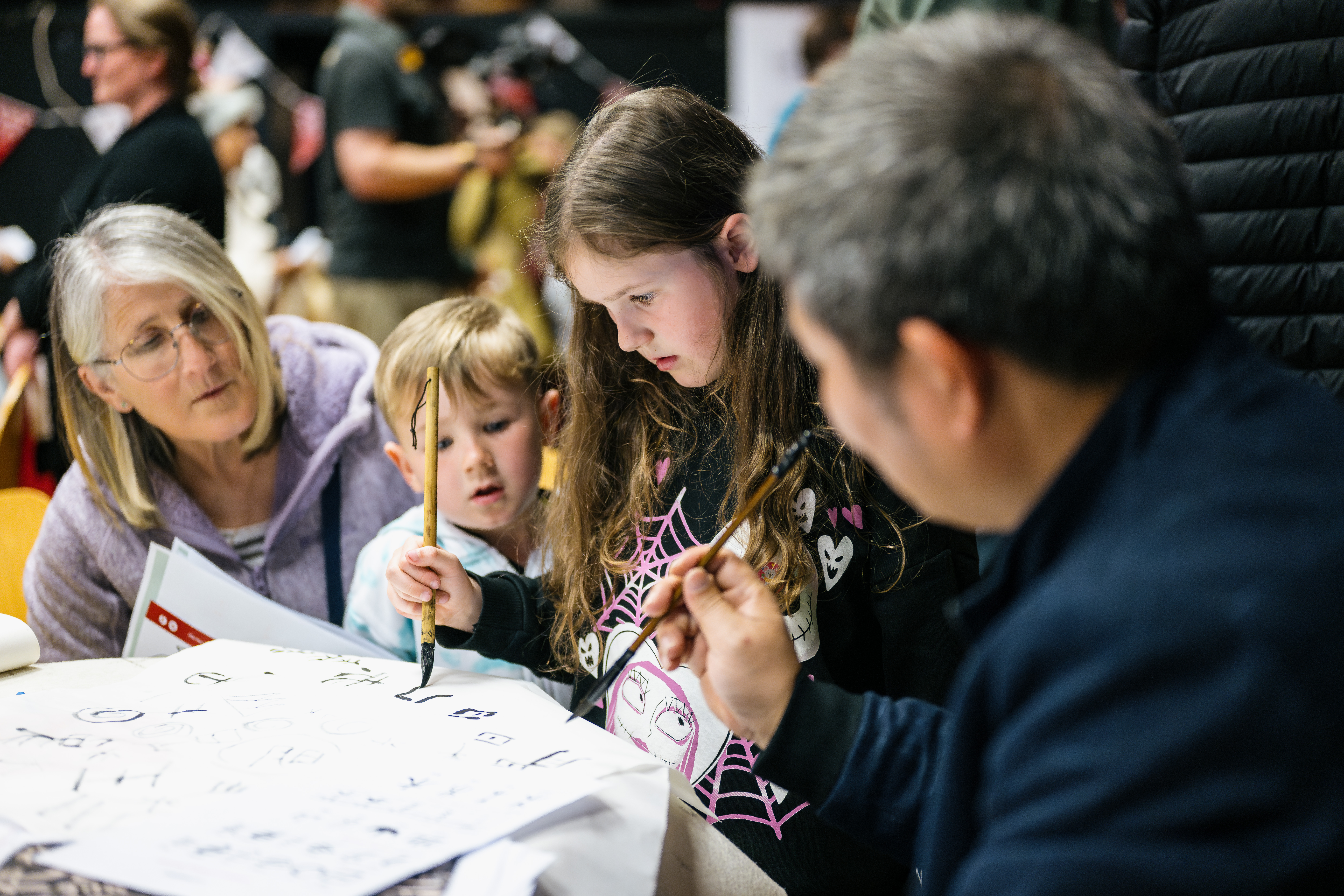 A young girl holds a calligraphy bursh whilst a small boy and 2 adults look on.