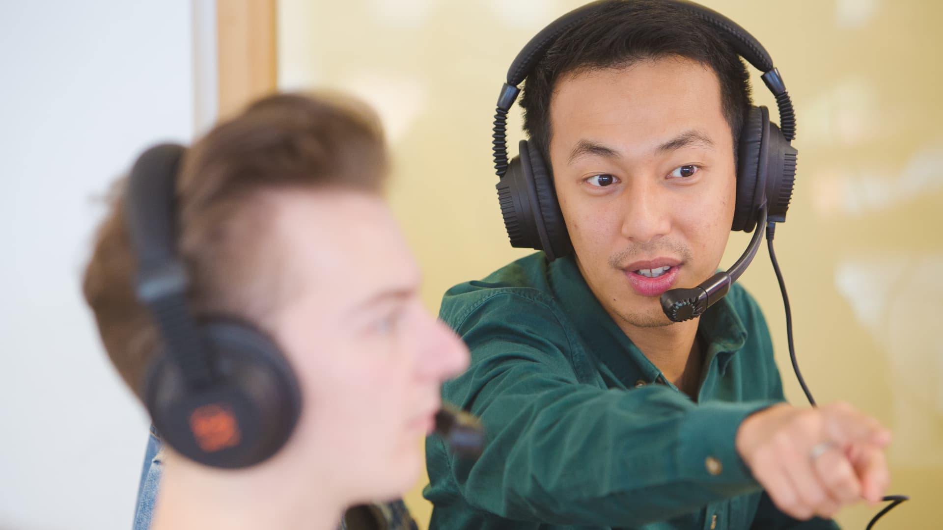 Two people wearing headsets are in a classroom. One is pointing at a screen to show the other person.