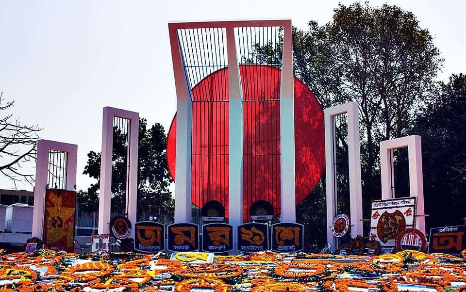 A tall white structure of pillars flanked by two smaller pillars and then two smaller pillars. Behind the pillars is a large red circle. On the ground are flower arrangements.