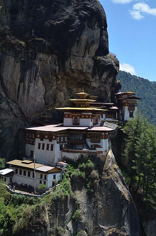  A Buddhist monastery perched in the mountains, the monastery has flat golden roofs and white walls. The mountain has a lot exposed rocks and trees near the base of the monastery.