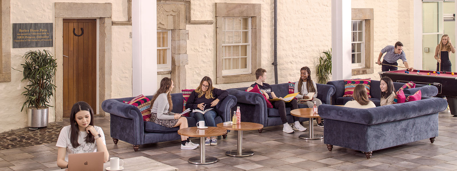 Several students sitting and chatting on sofas while two in the background play pool.