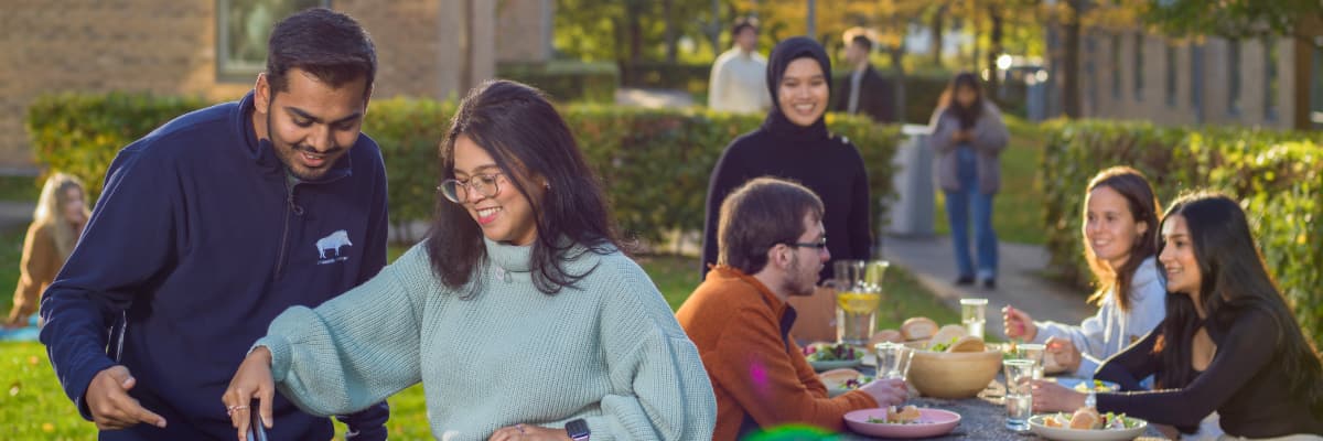 Students operate a barbecue, whilst more sit at a picnic table. In the background are townhouses.