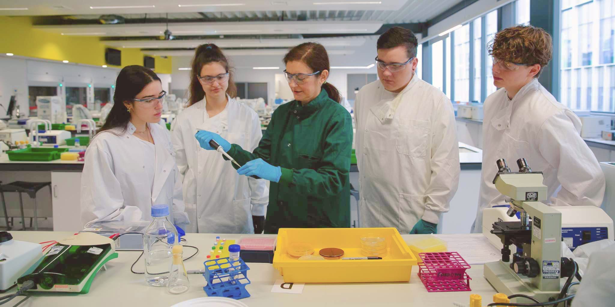 Students in white lab coats in the lab with an lecturer wearing a green lab coat demonstrating