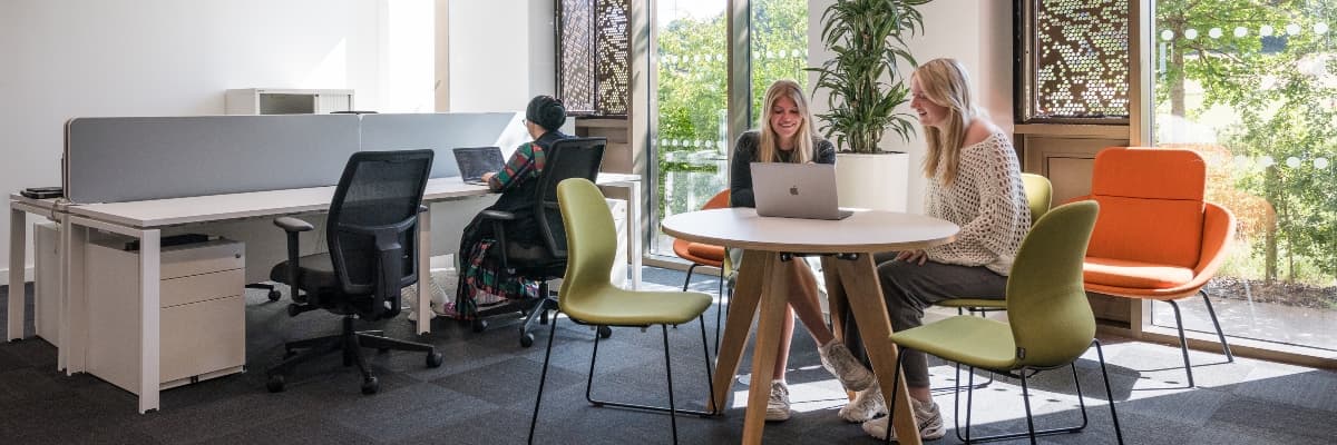 Three people working with laptops in an office, one at a desk and two seated at a coffee table