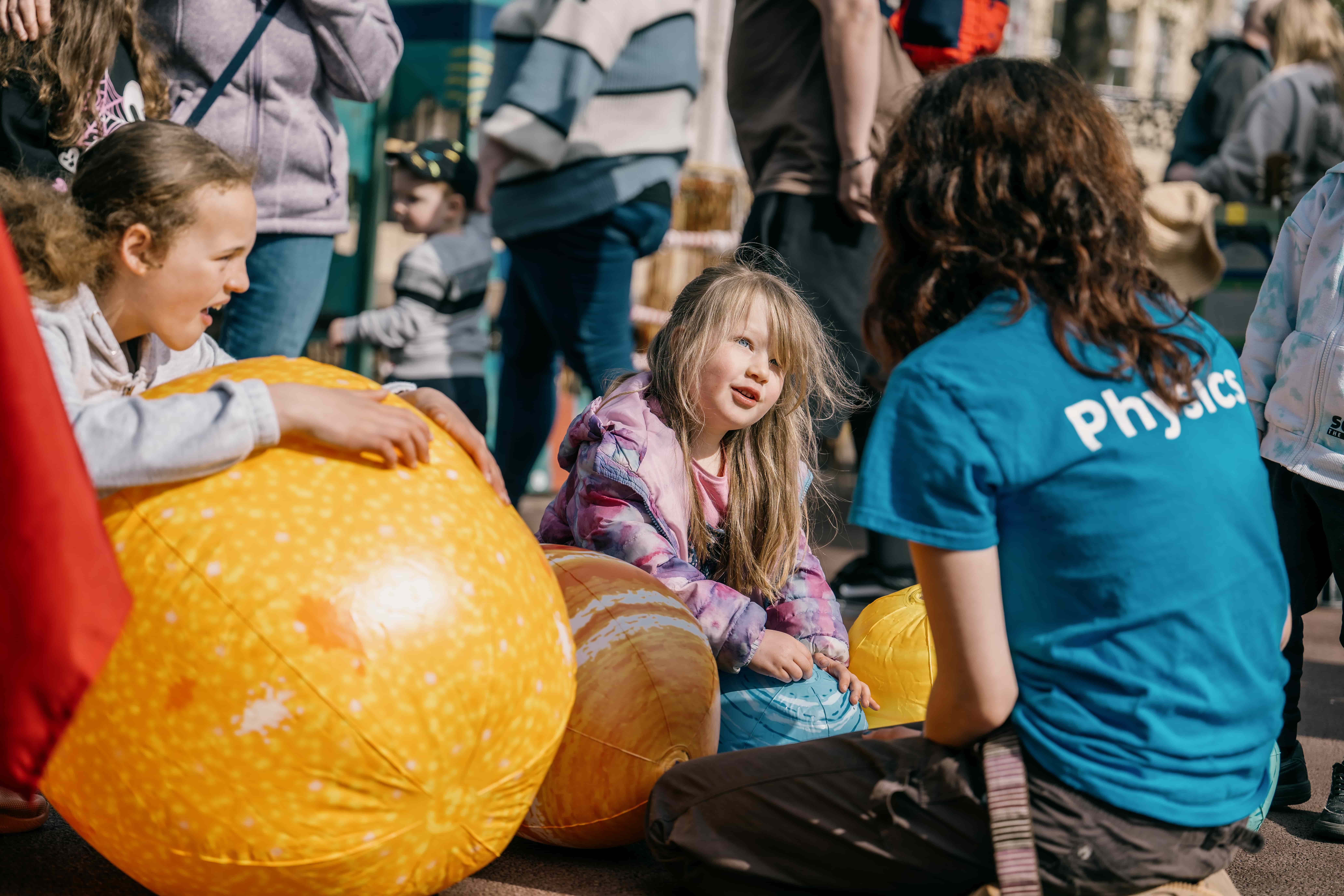 2 girls sit holding on to inflable planets listening to a student in a tshirt labelled physics