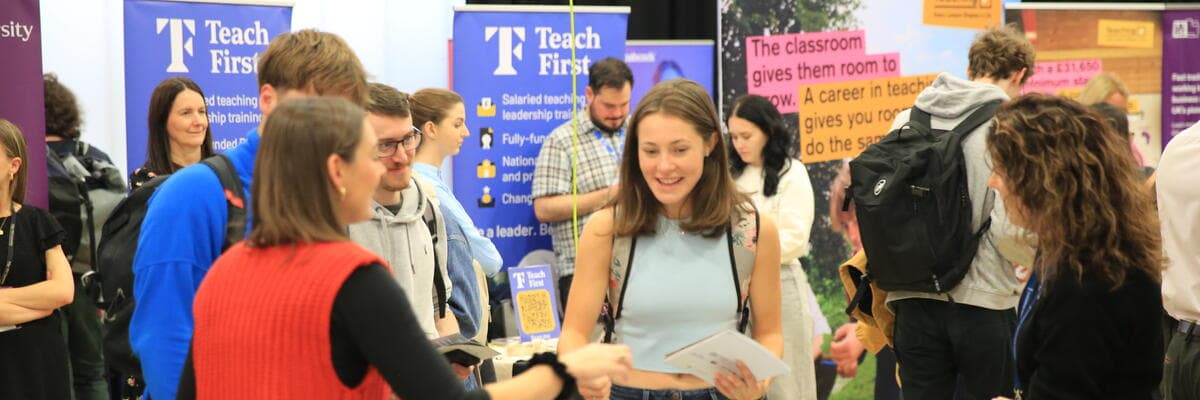 Students speaking with employers at a busy careers fair, with employer banners in the background.