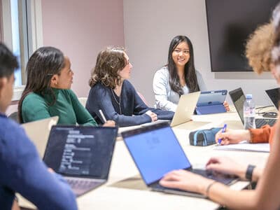 Students work together around a table in a meeting room, using laptops during a group discussion.