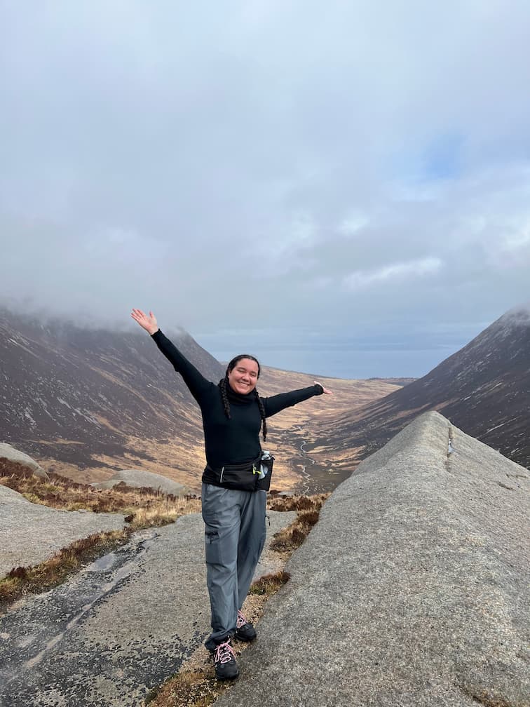 Image of student Lyea with arms out smiling atop a mountain