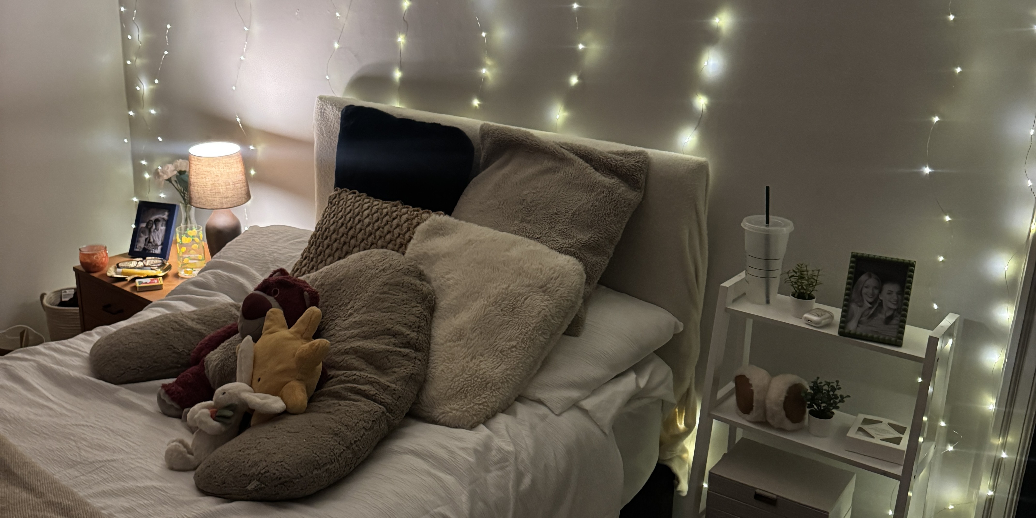 Cozy bedroom featuring a string of fairy lights along the wall, fluffy pillows, a stuffed toy, and a blanket on the bed. The bedside areas are decorated with photos, candles, and indoor plants.