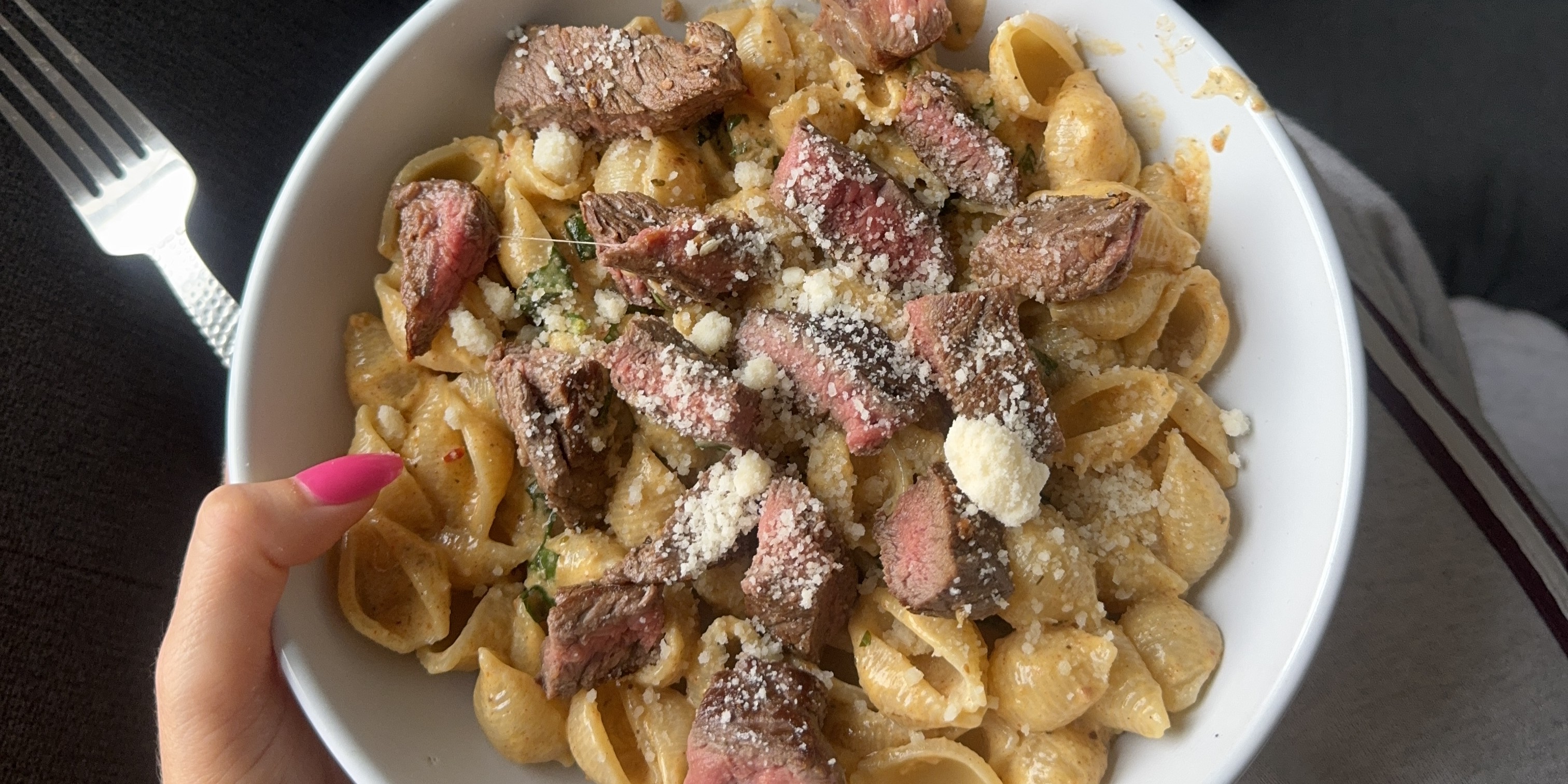 A person holding a bowl of steak pasta sprinkled with cheese on a cozy couch.