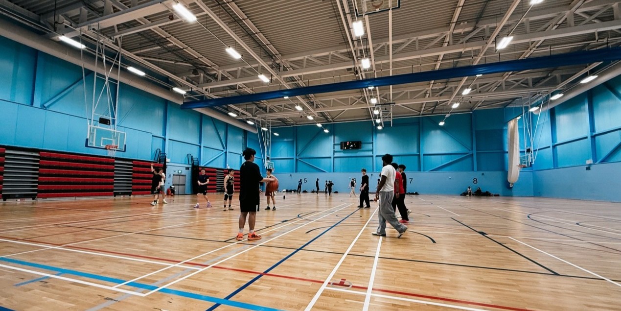A group of people playing basketball in the basketball court at sports centre.