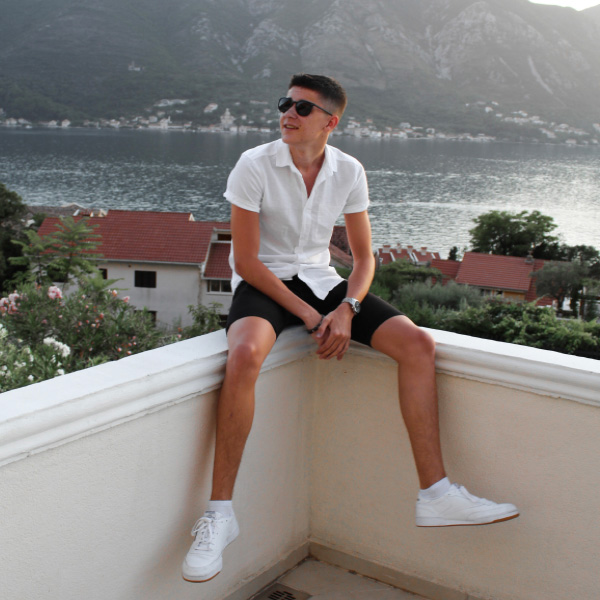 A male student Wearing sunglasses, shorts and a shirt, sitting on a wall overlooking the sea.