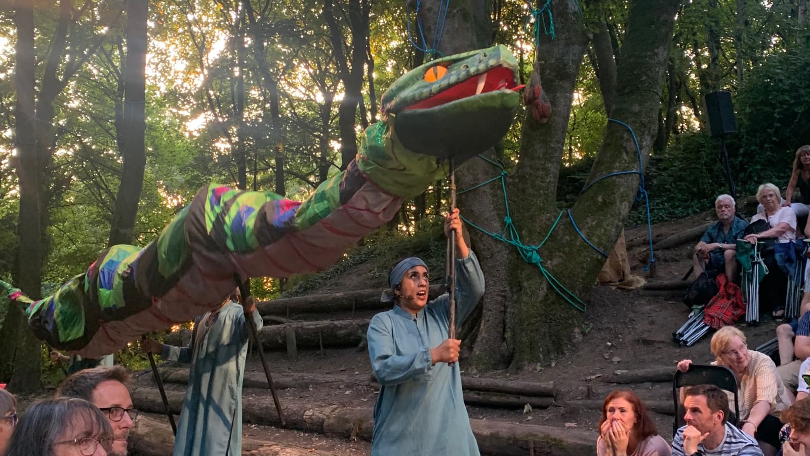 Actors manipulate a giant snake puppet in a wooded glade.