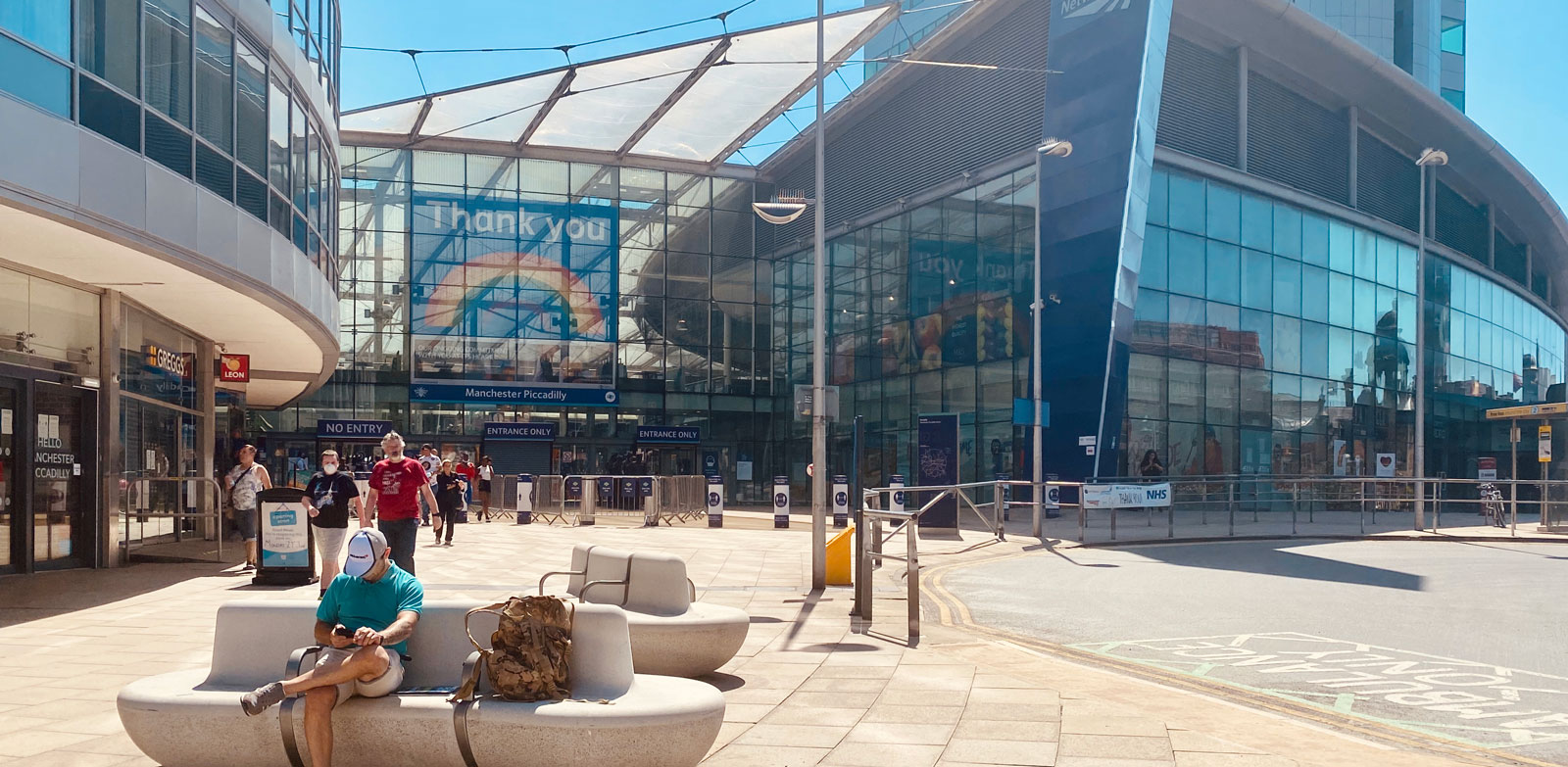 Manchester Picadilly station entrance on a sunny day. A few people are sat around or walking.