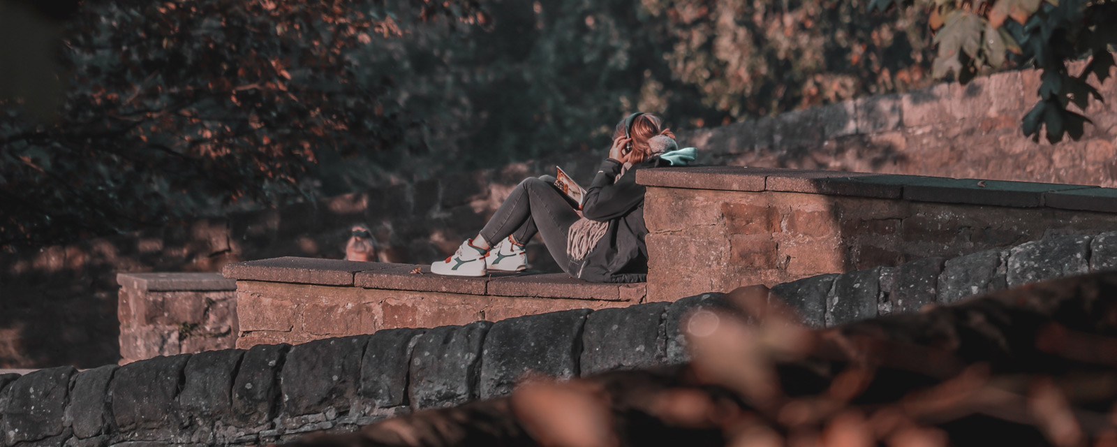 A female student sitting on a wall reading and wearing headphones
