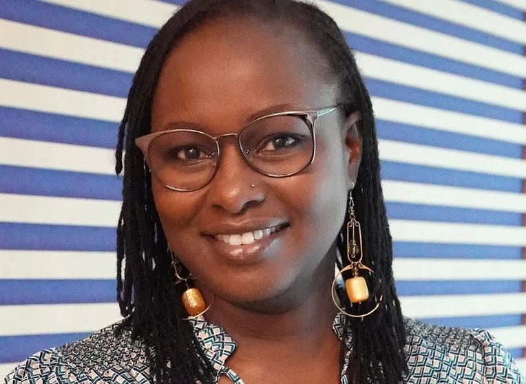 Moderator Kendi Guantai smiles at the camera while standing in front of a striped backdrop.