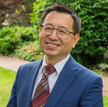 Yanguo stands outside in front of a hedge. He wears a blue suit and dark red tie.