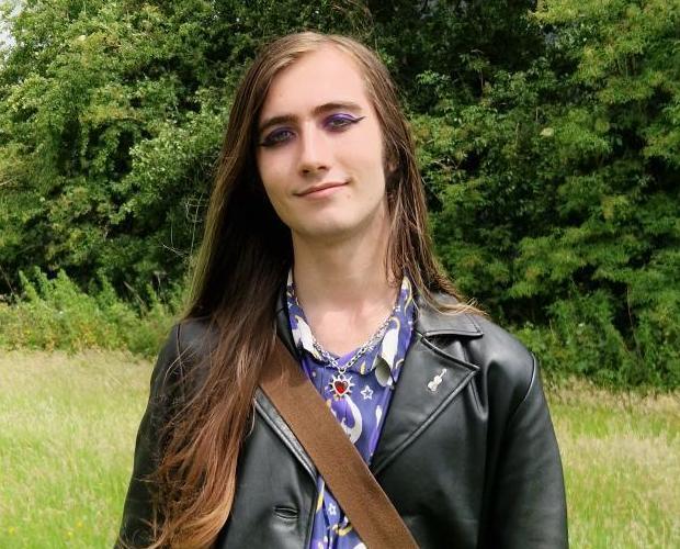 Siven stands in a green field, wearing a purple shirt and black jacket looking towards the camera