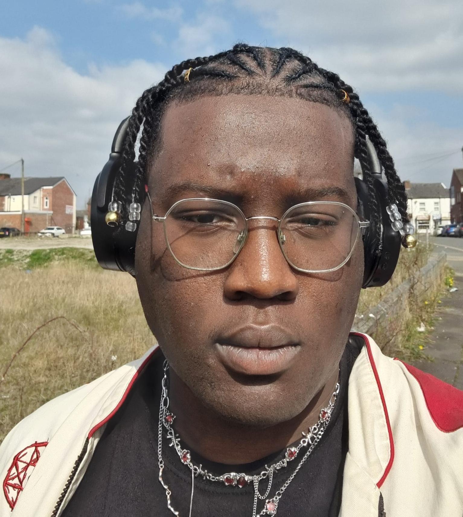 Uyi stands outside. He is wearing headphones, a cream jacket, and glasses
