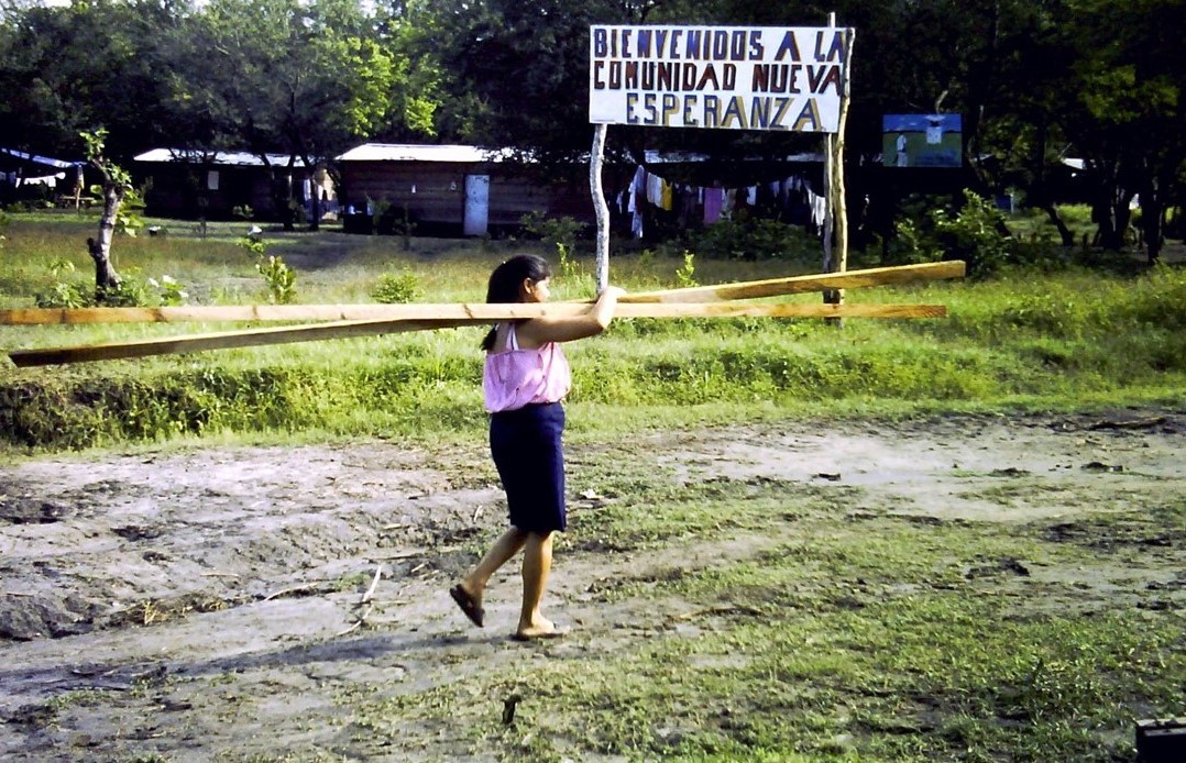 A young woman carrying wood in front of a sign reading: Bienvenidos a la comunidad Nueva Esperanza.