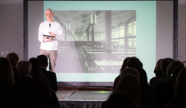 Joe Moran stands in front of a screen showing an image of a refectory. 