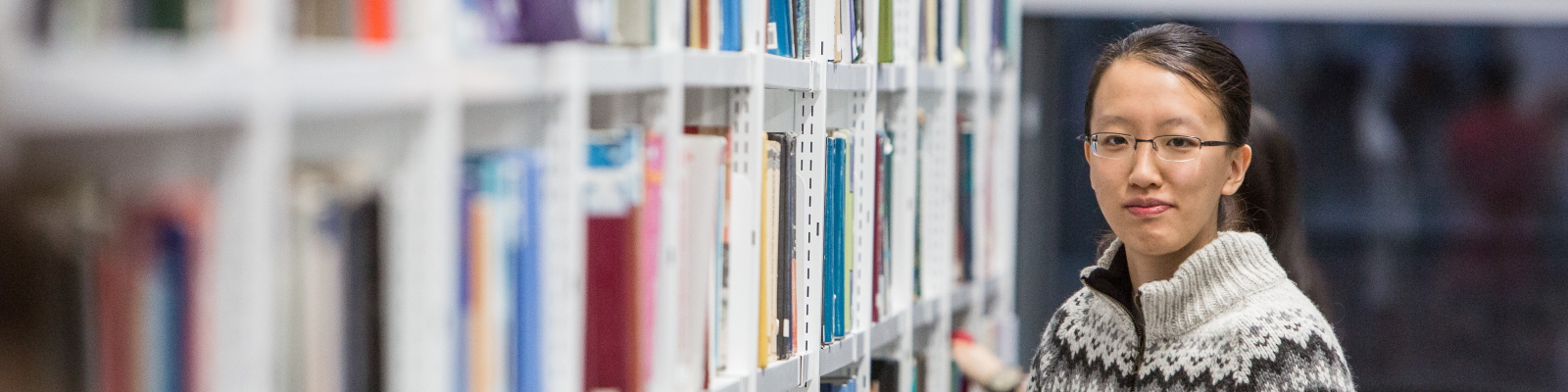 A student standing next to shelves containing books, in the Library.