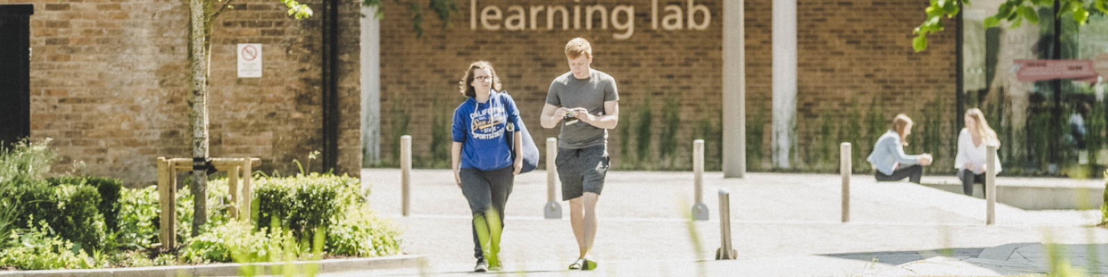 Two students walking across campus, with buildings behind and grass and trees in the foreground.