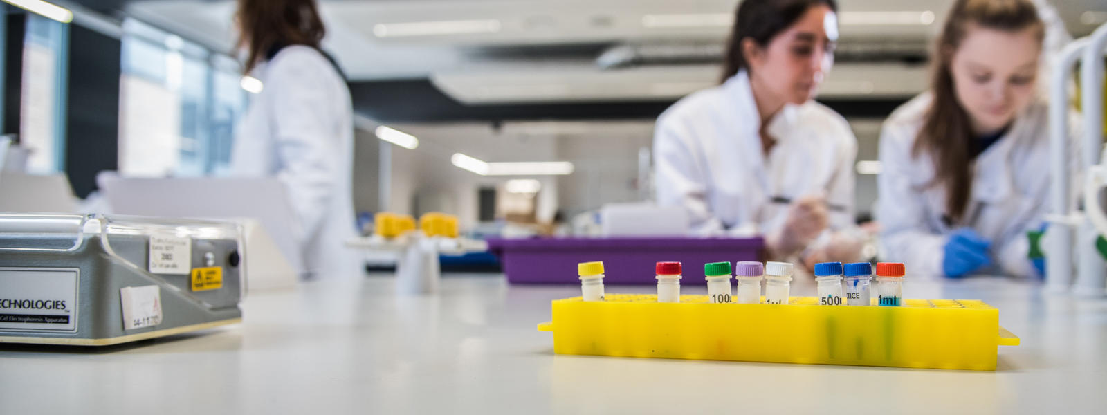 A close up of some liquid vials on the laboratory work-surface, with students working in the background.