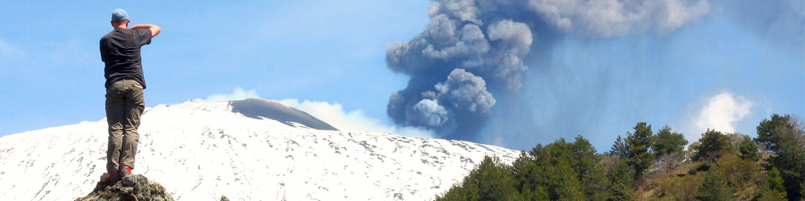 A man stands on a rocky outcrop photographing an eruption on Mount Etna