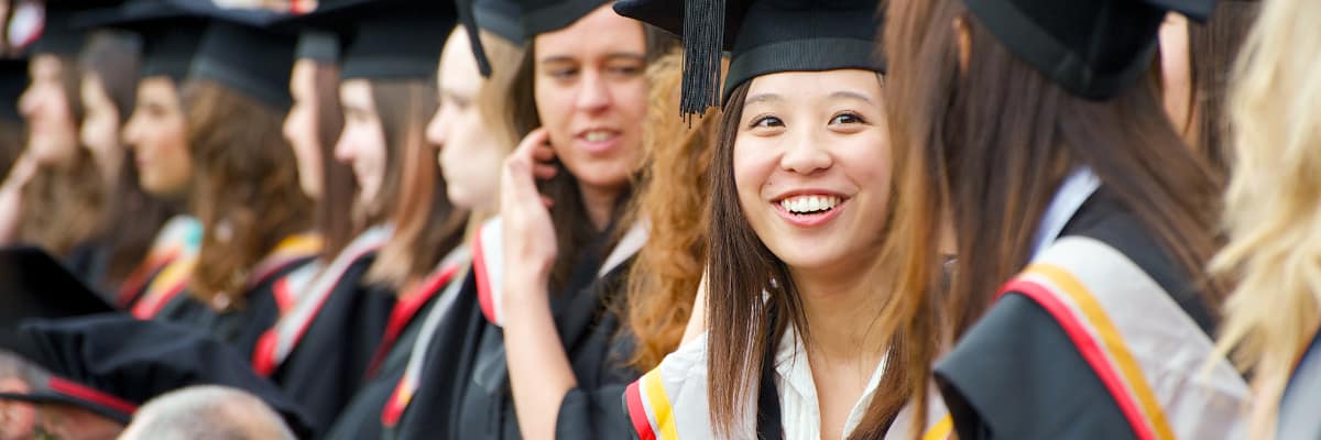 A student in graduation robes smiles, whilst standing among a crowd.