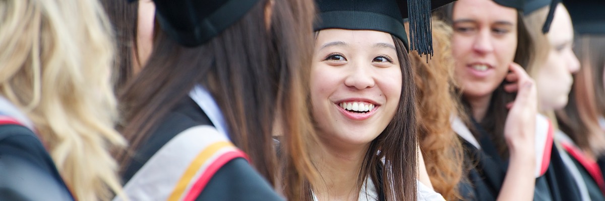 A student in graduation robes smiles, whilst standing amongst a crowd.