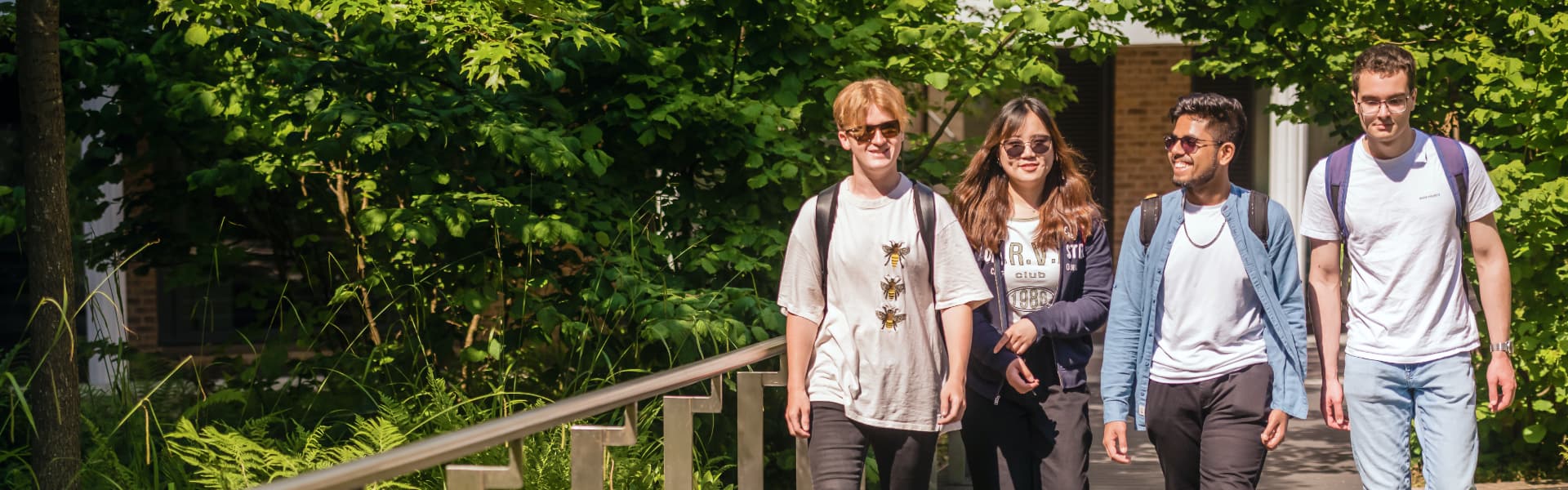 4 students walk across the campus, surrounded by greenery. It is very sunny.