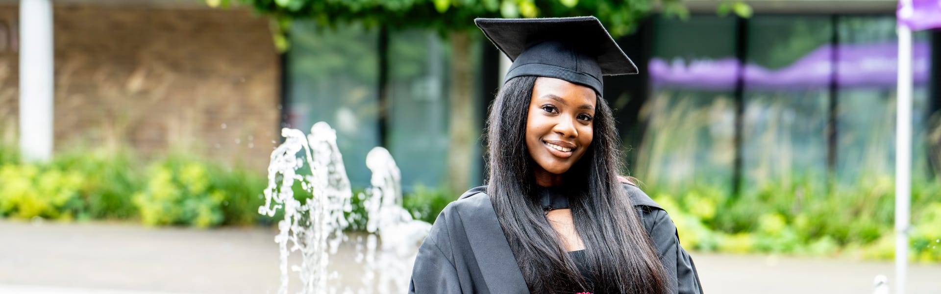 A smiling student stands in graduation gowns in front of a fountain