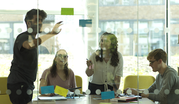 Students in a study space putting sticky notes on a glass wall.