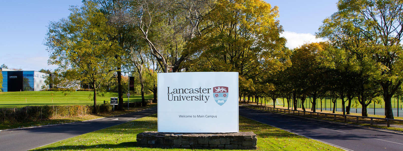 Entrance road into Lancaster campus. A sign surrounded by trees says welcome.