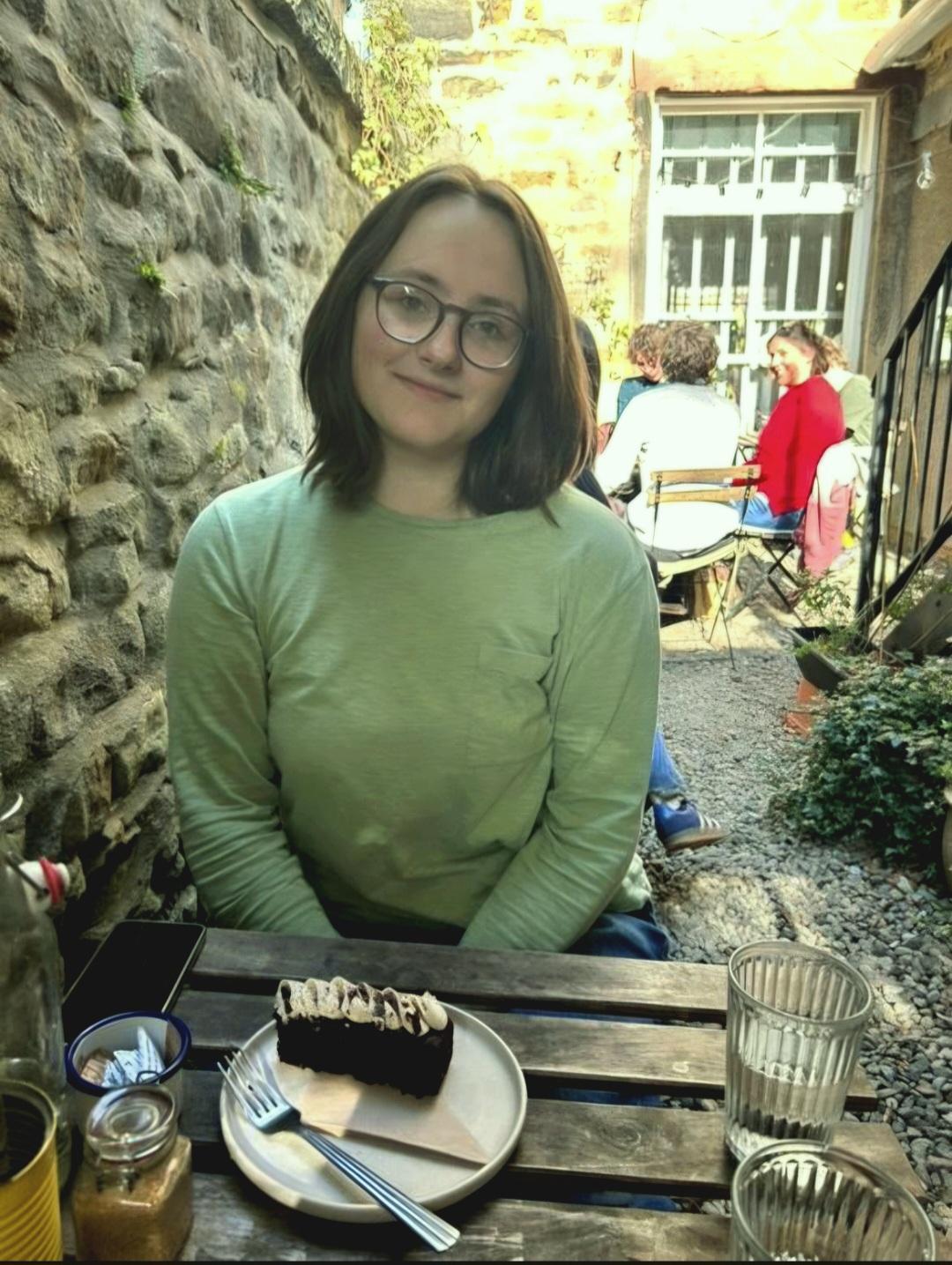 Luna smiling at the camera, wearing glasses and a green top, seated at an outdoor café table with a 