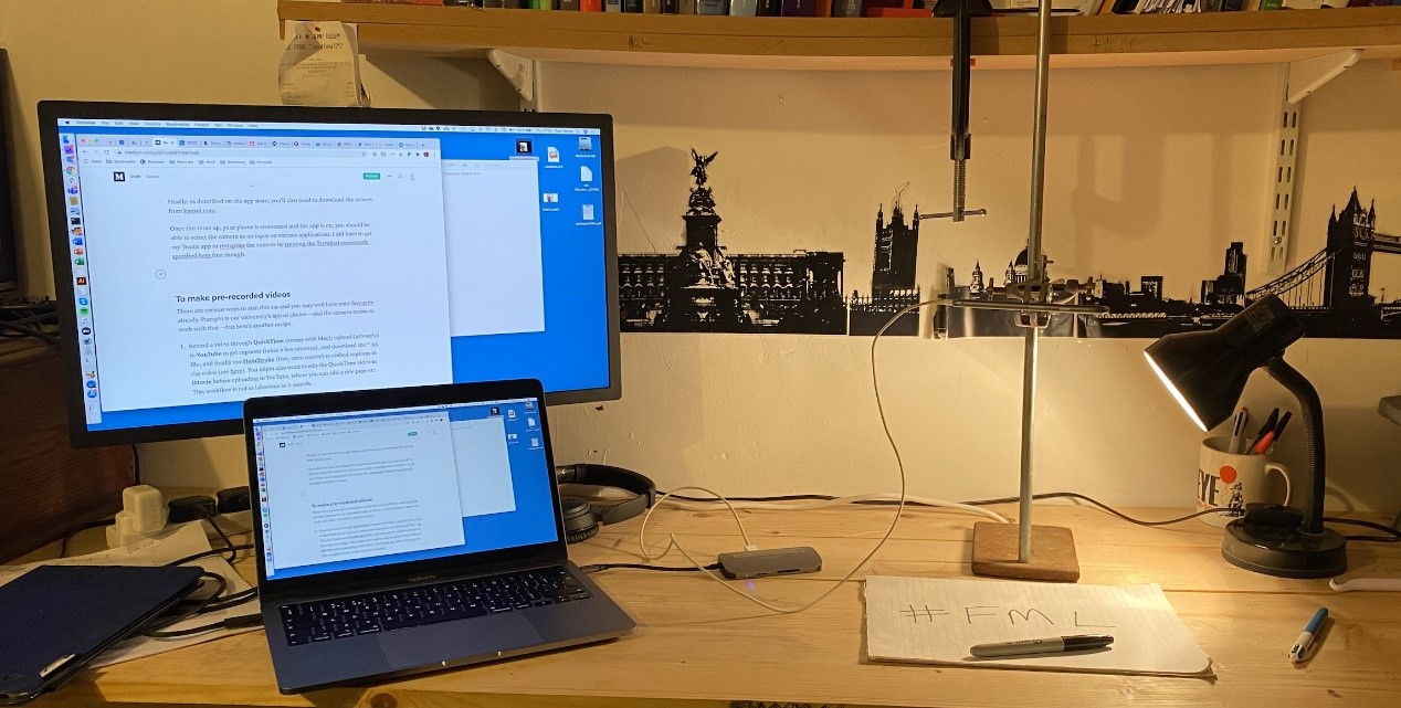 A desk with a laptop and monitor and a homemade visualiser lit by a lamp.