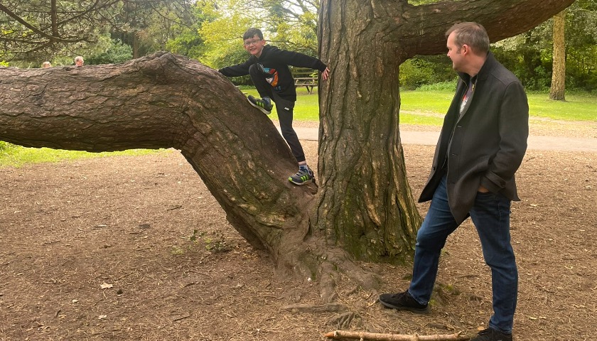 A child climbs in a tree whilst a parent watches on, smiling.