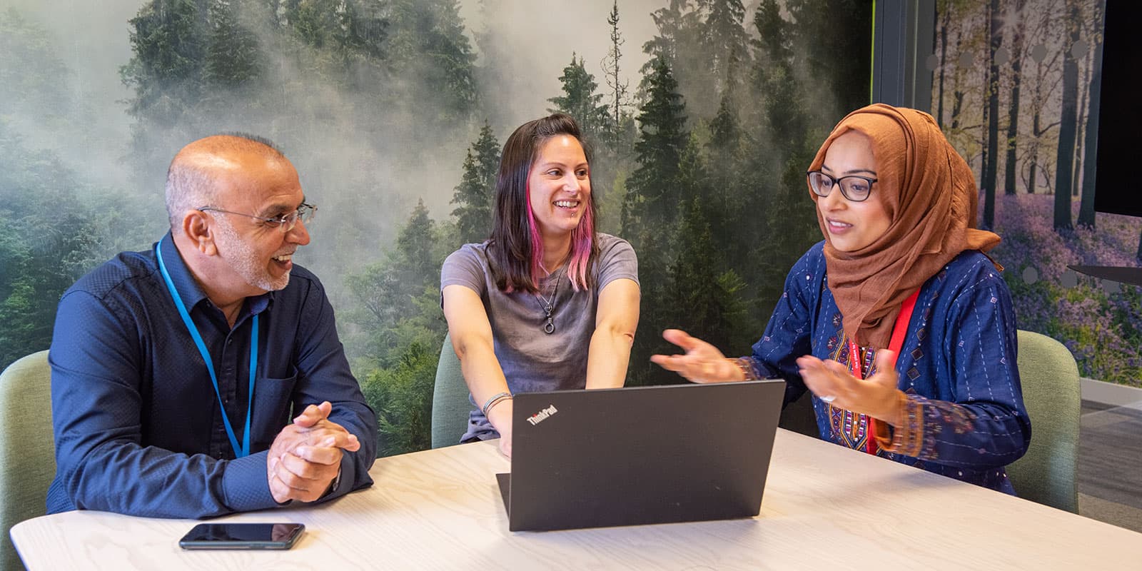 Three people sit around a laptop computer in a modern office space.