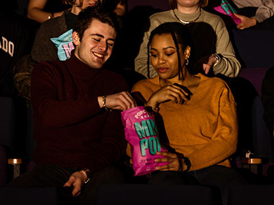 Two people siting in the cinema sharing a large packet of crisps.
