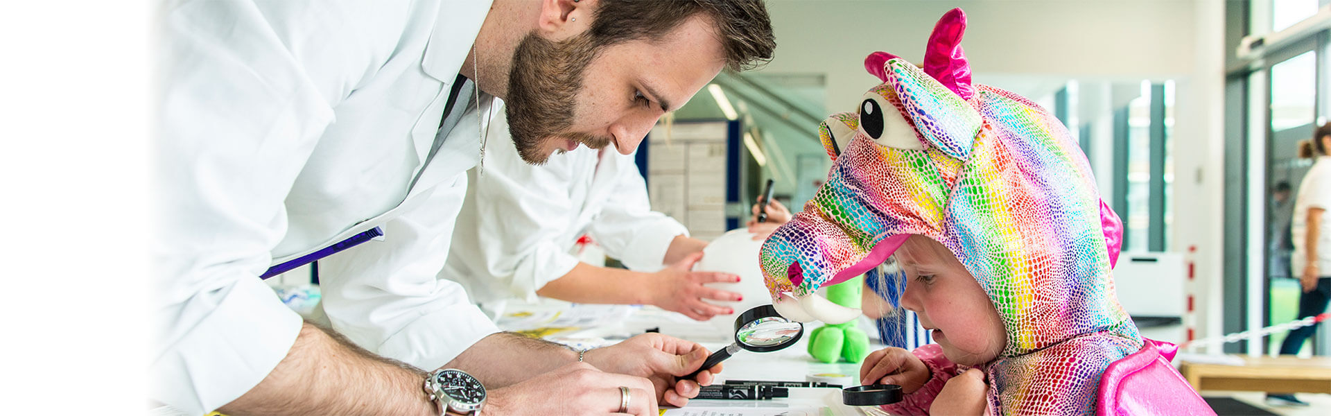 A university scientist demonstrates a experiment to a young girl in a multi-coloured dragon costume