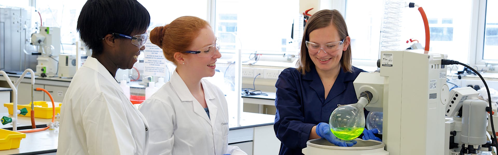 Two students in a chemistry laboratory talk to a laboratory assistant.