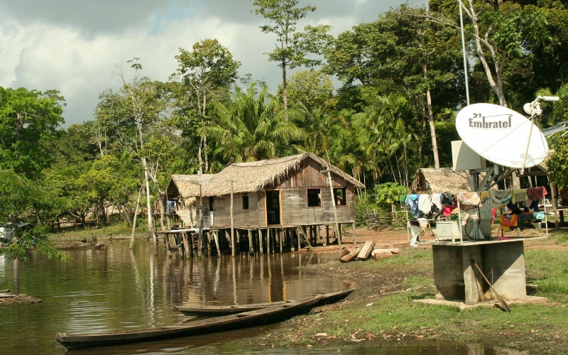 Beside a tropical river sits a wooden house on stilts with a large satellite dish outside