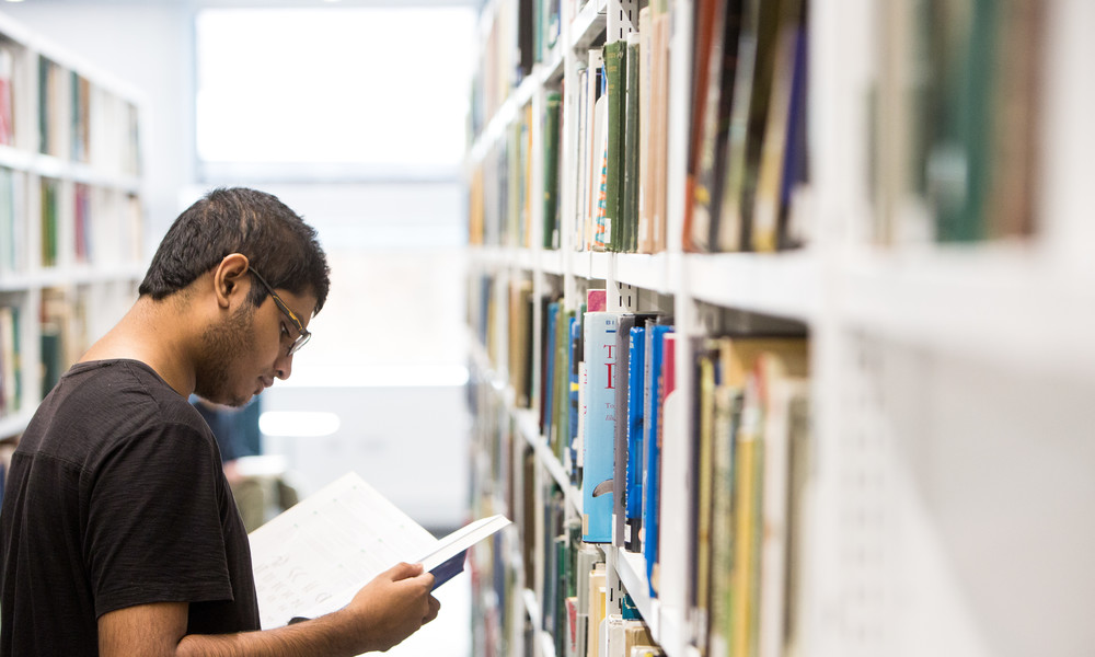 Student reading a book next to a bookshelf.