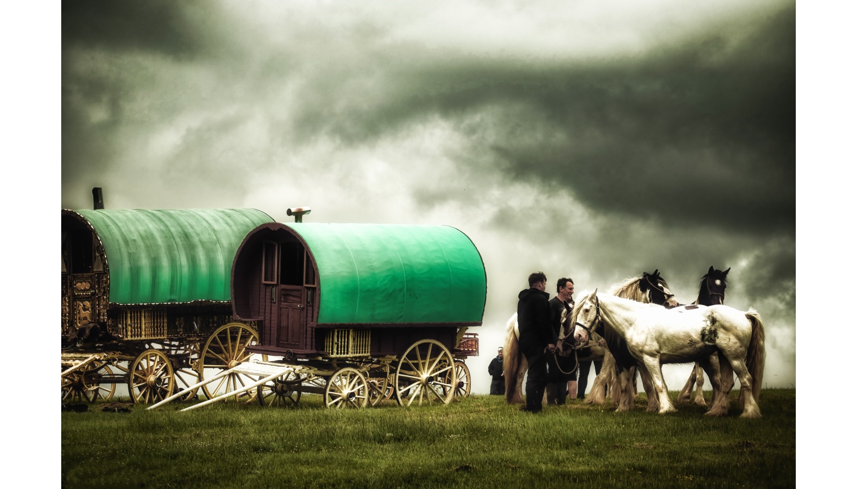 Two men standing next to three horses and two canvas covered caravans.