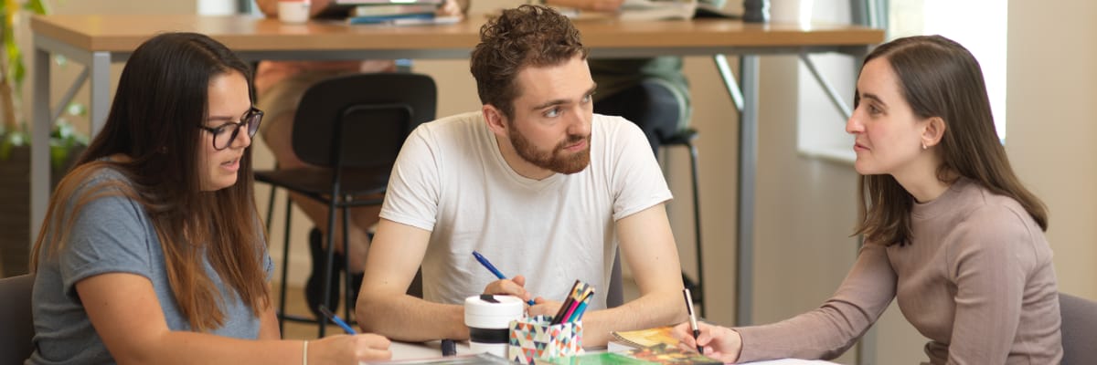 Three students sit around a table writing and talking.
