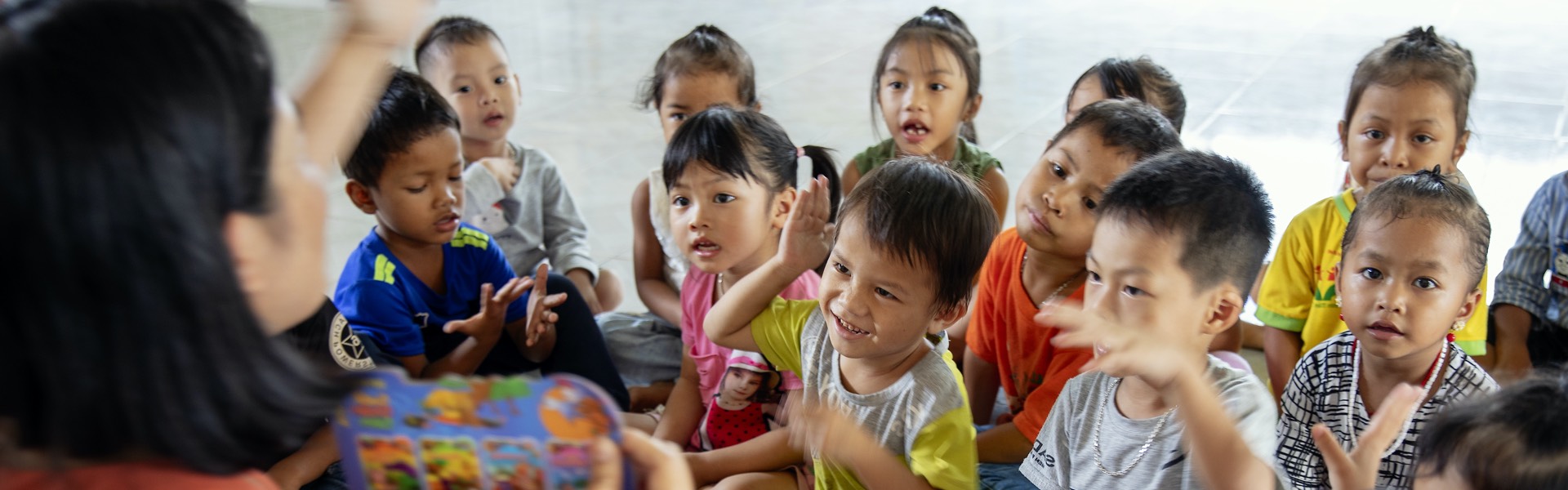 Photograph of a class of children sat on a carpet listening to a teacher reading in Viet Nam.